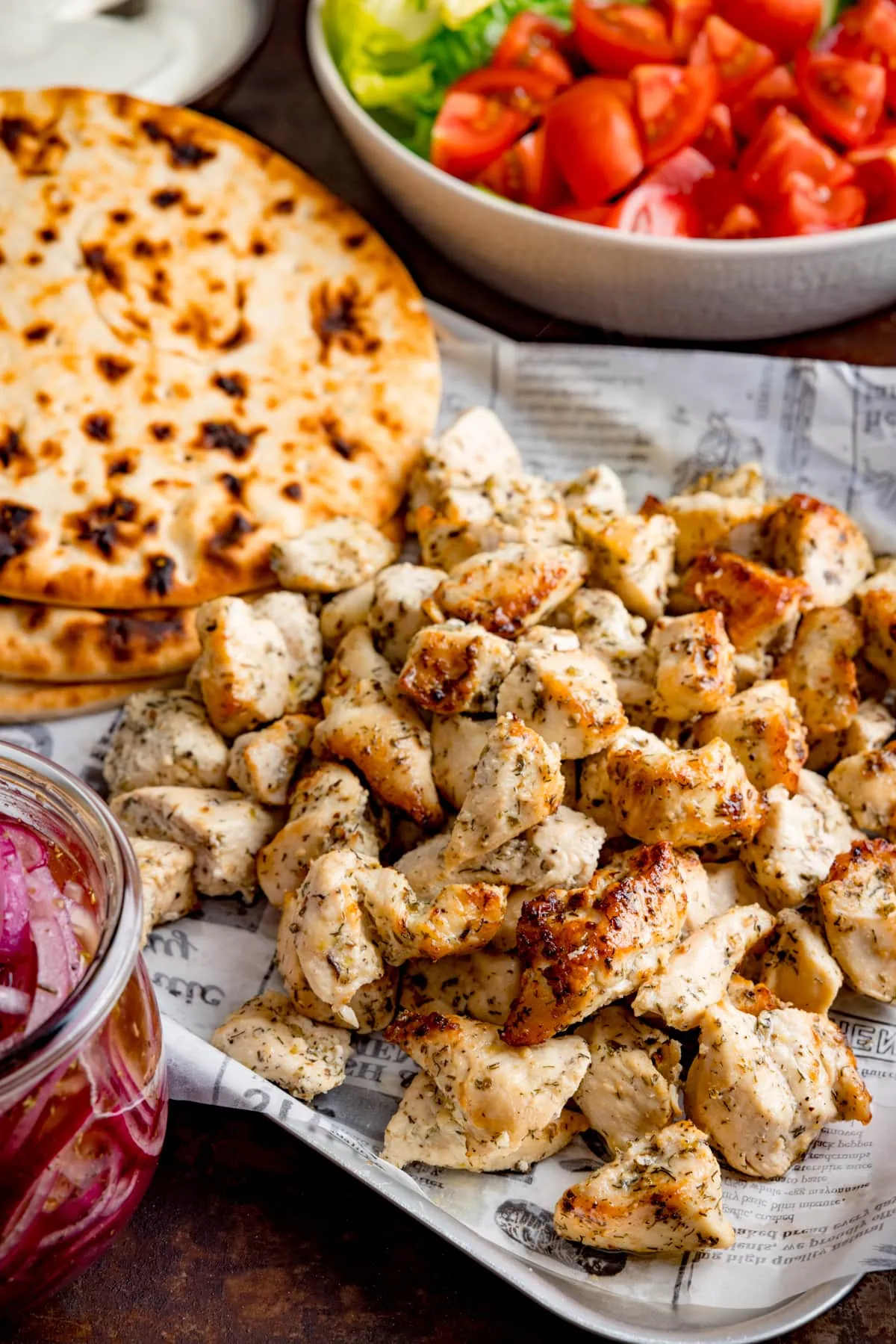 Air fryer greek chicken pieces on a tray with flatbreads, a bowl of tomatoes and lettuce, and a jar of marinated onions.