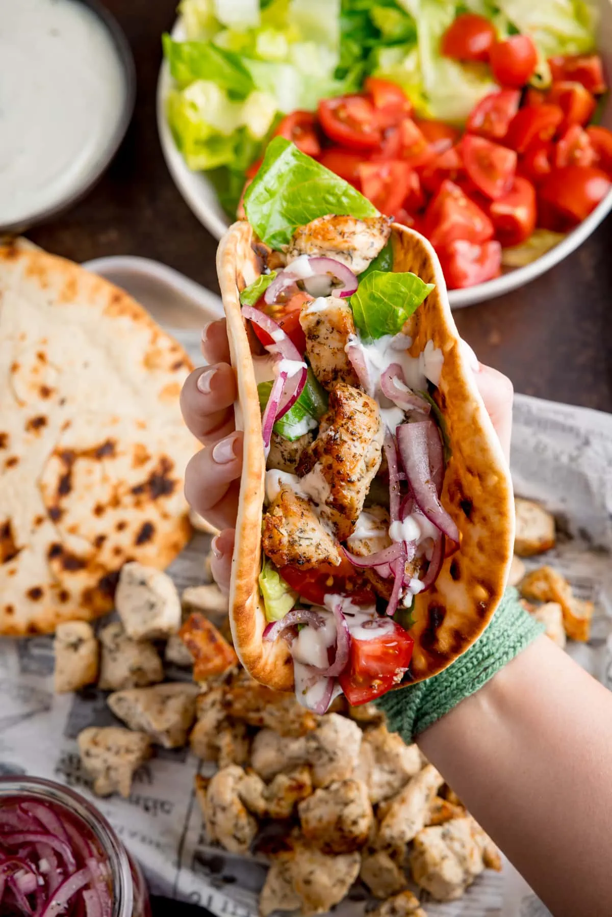 Overhead shot of air fryer Greek chicken pieces in a flatbread with salad and feta dressing. The wrap is being held by a hand over the top of a tray that has all of the cooked ingredient on it.