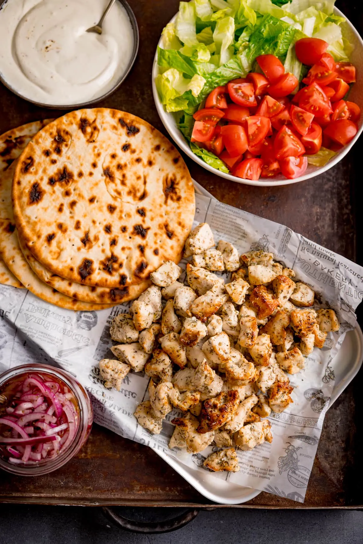 Overhead image of air fryer greek chicken pieces on a tray with flatbreads, a bowl of tomatoes and lettuce, a jar of marinated onions and a bowl of cream feta dip.