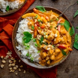 Square overhead image of chicken panang curry and rice in a brown bowl, topped with chopped peanuts and Thai basil leaves. There is a further bowl, just in shot, at the top of the frame, an orange napkin next to the bowls and some chopped peanuts nearby. The bowls are on a wooden table.