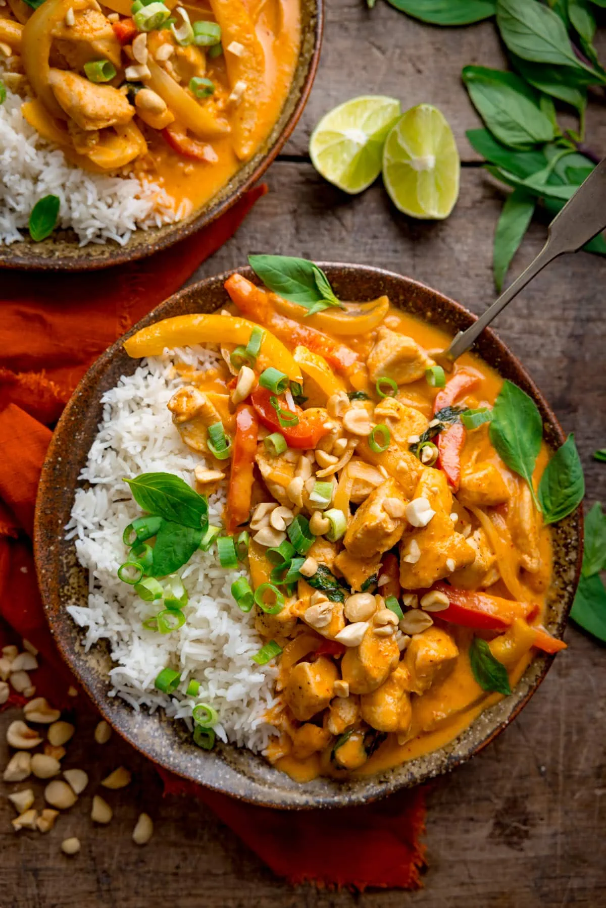 Overhead tall shot of chicken panang curry and rice in a brown bowl, topped with chopped peanuts and Thai basil leaves. There is a further bowl at the top of the frame, an orange napkin next to the bowls and some chopped peanuts, Thai basil leaves and limes wedges nearby. The bowls are on a wooden table.