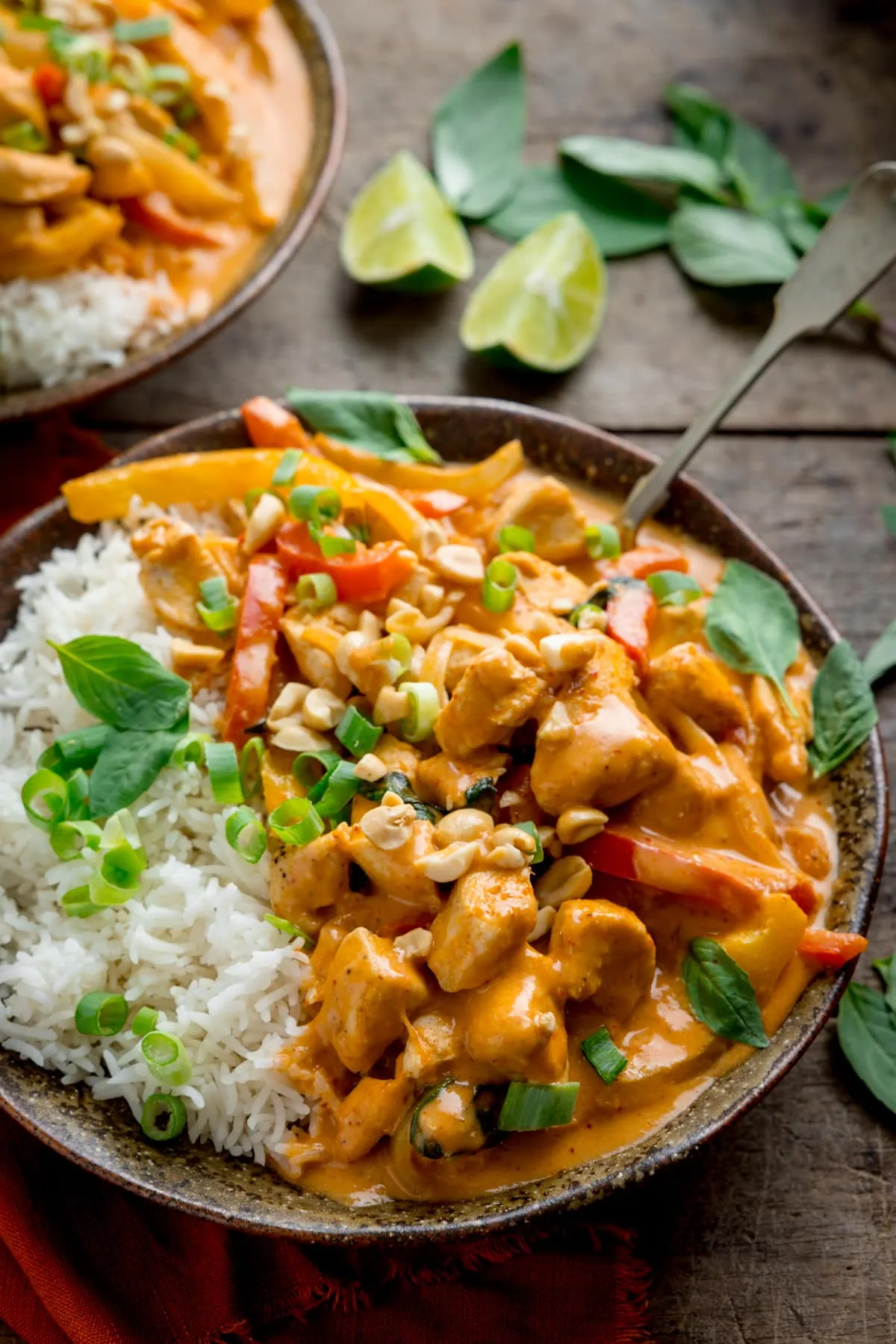 Side on shot of chicken panang curry and rice in a brown bowl, topped with chopped peanuts and Thai basil leaves. There is a further bowl at the top of the frame and some Thai basil leaves and limes wedges nearby. The bowls are on a wooden table.