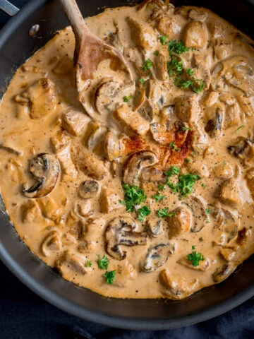 Square overhead image of chicken and mushroom stroganoff in a dark pan. The pan is on a dark surface with a wooden spoon sticking out. The stroganoff is garnished with chopped parsley and paprika.