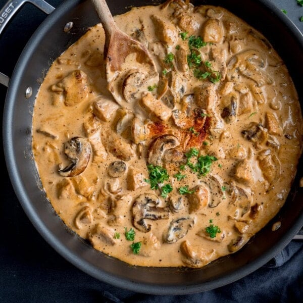 Square overhead image of chicken and mushroom stroganoff in a dark pan. The pan is on a dark surface with a wooden spoon sticking out. The stroganoff is garnished with chopped parsley and paprika.