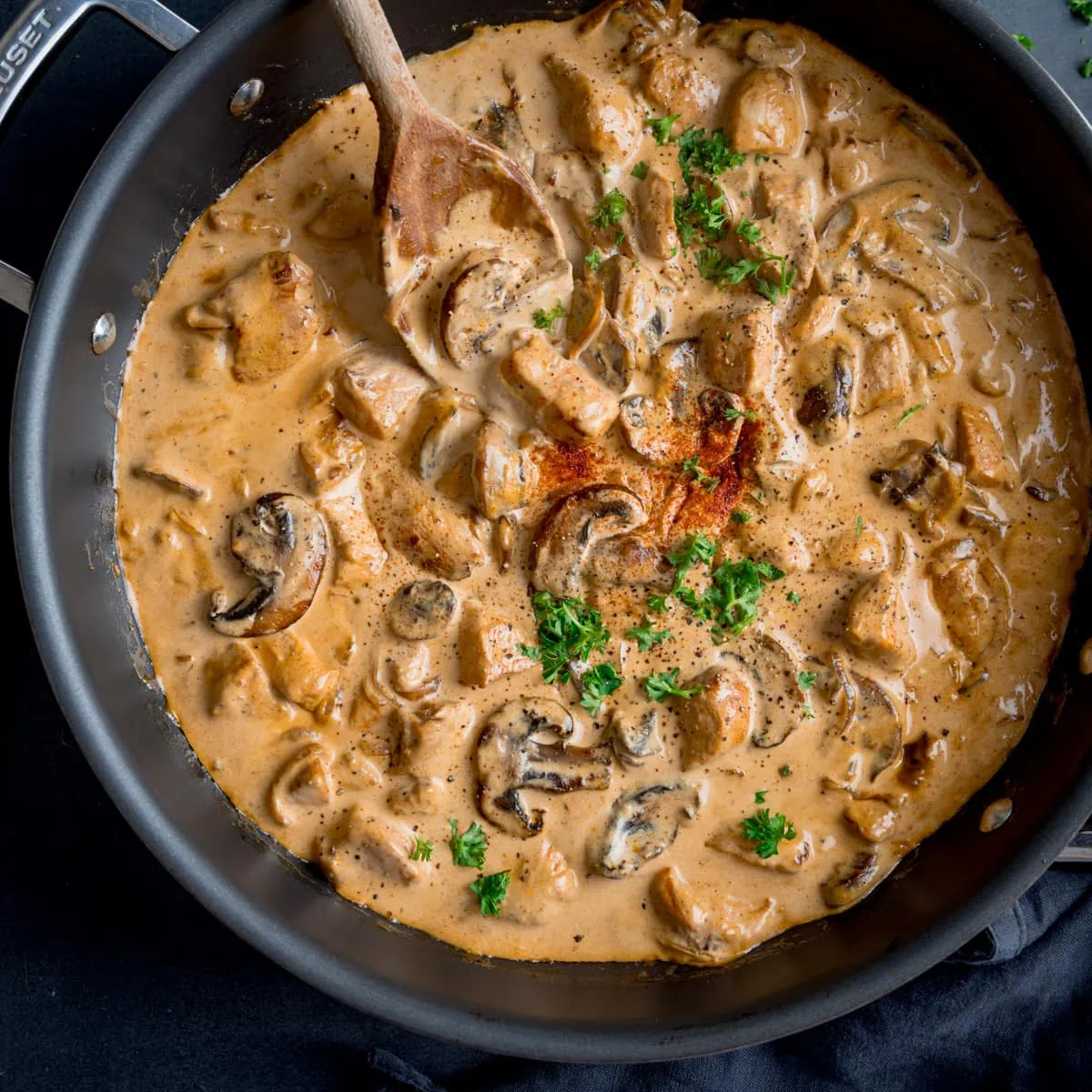 Square overhead image of chicken and mushroom stroganoff in a dark pan. The pan is on a dark surface with a wooden spoon sticking out. The stroganoff is garnished with chopped parsley and paprika.