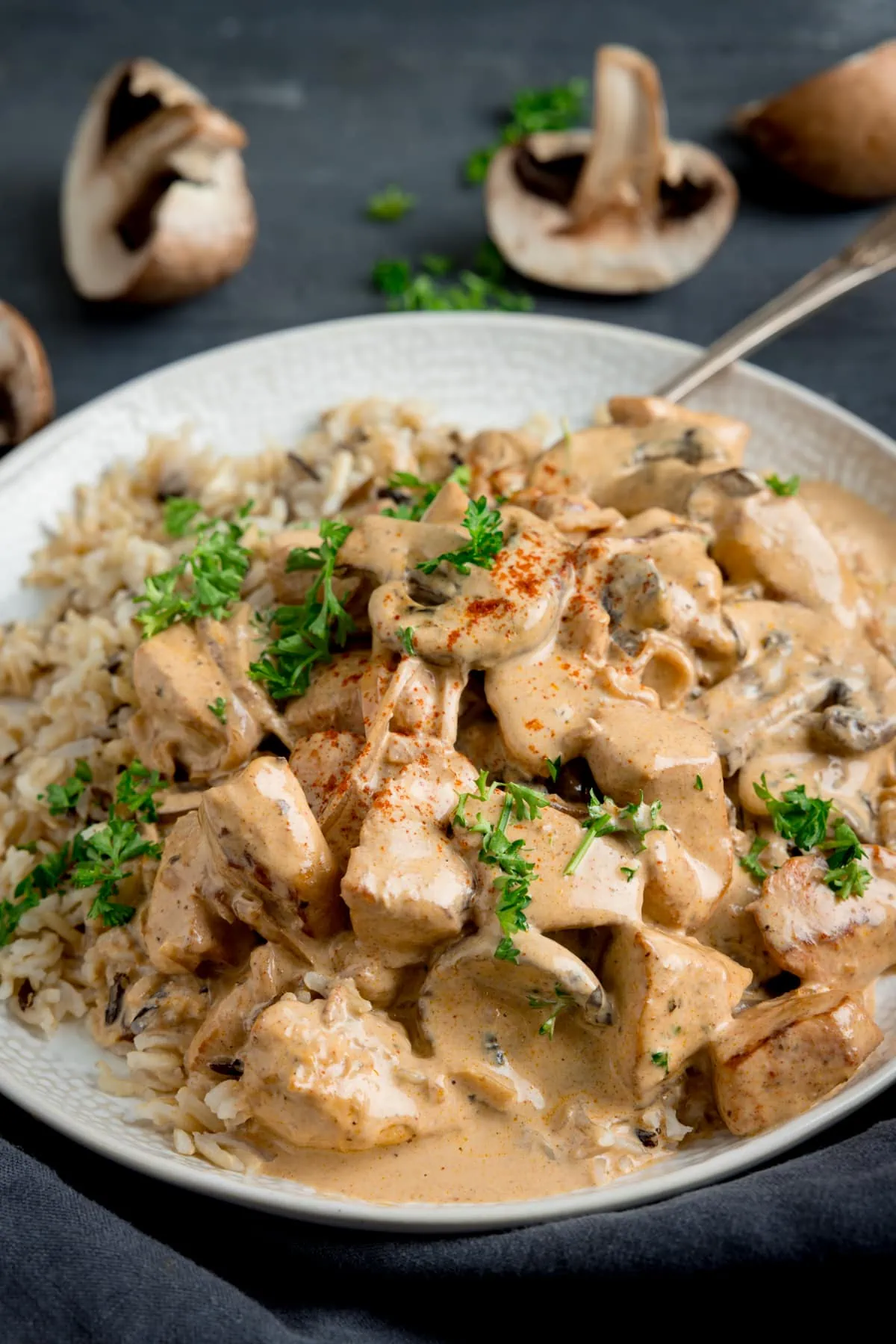 Side-on image of chicken stroganoff and rice on a white plate with a metal fork. The plate is on a dark surface and there are sliced mushrooms and chopped parsley scattered around.