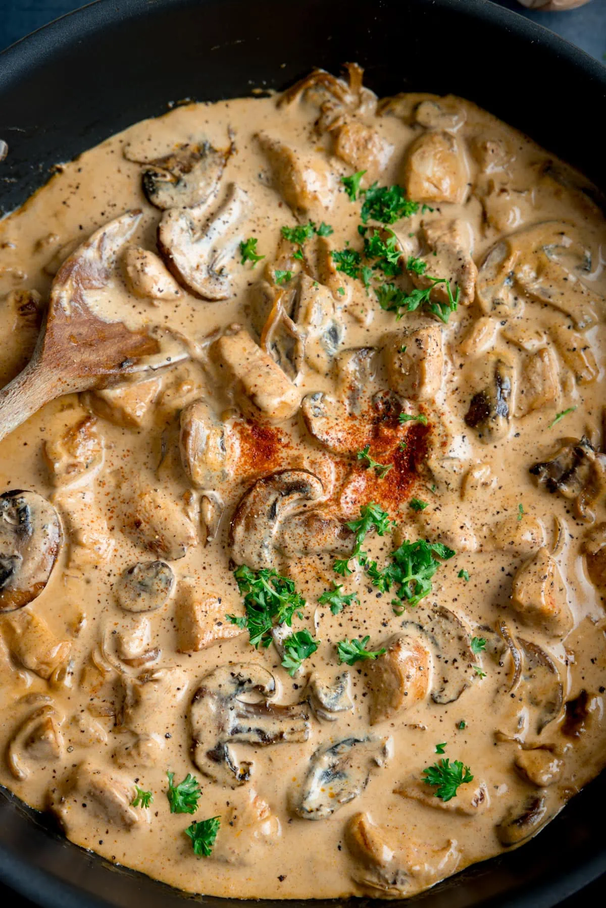 Tall, overhead, close-up image of chicken and mushroom stroganoff in a dark pan. The pan is on a dark surface with a wooden spoon sticking out. The stroganoff is garnished with chopped parsley and paprika.