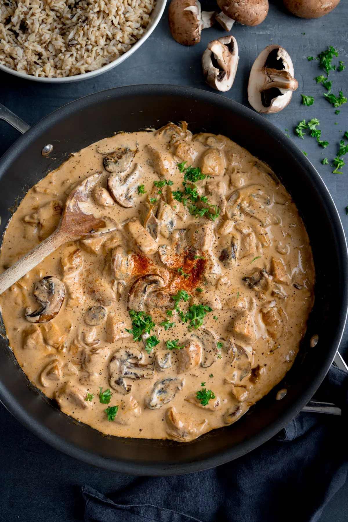 Tall overhead shot of chicken and mushroom stroganoff in a dark pan. The pan is on a dark surface with a wooden spoon sticking out. There is a bowl of cooked rice next to the pan and some mushrooms and chopped parsley scattered around.