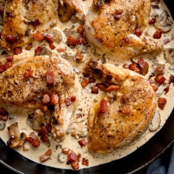 Overhead close-up square image of chicken supreme with mushrooms and pancetta in a black pan. There are four chicken supremes in the pan.