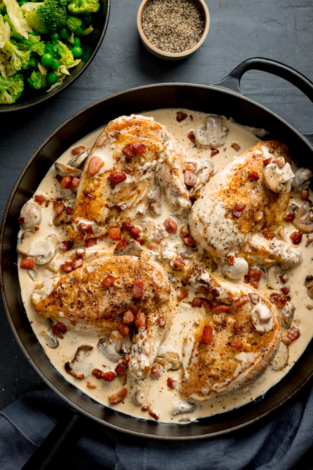 Tall overhead image of chicken supreme with mushrooms and pancetta in a black pan. There are four chicken supremes in the pan. The pan is on a dark background next to a dark napkin, a bowl of green veg and a pinch pot of black pepper.