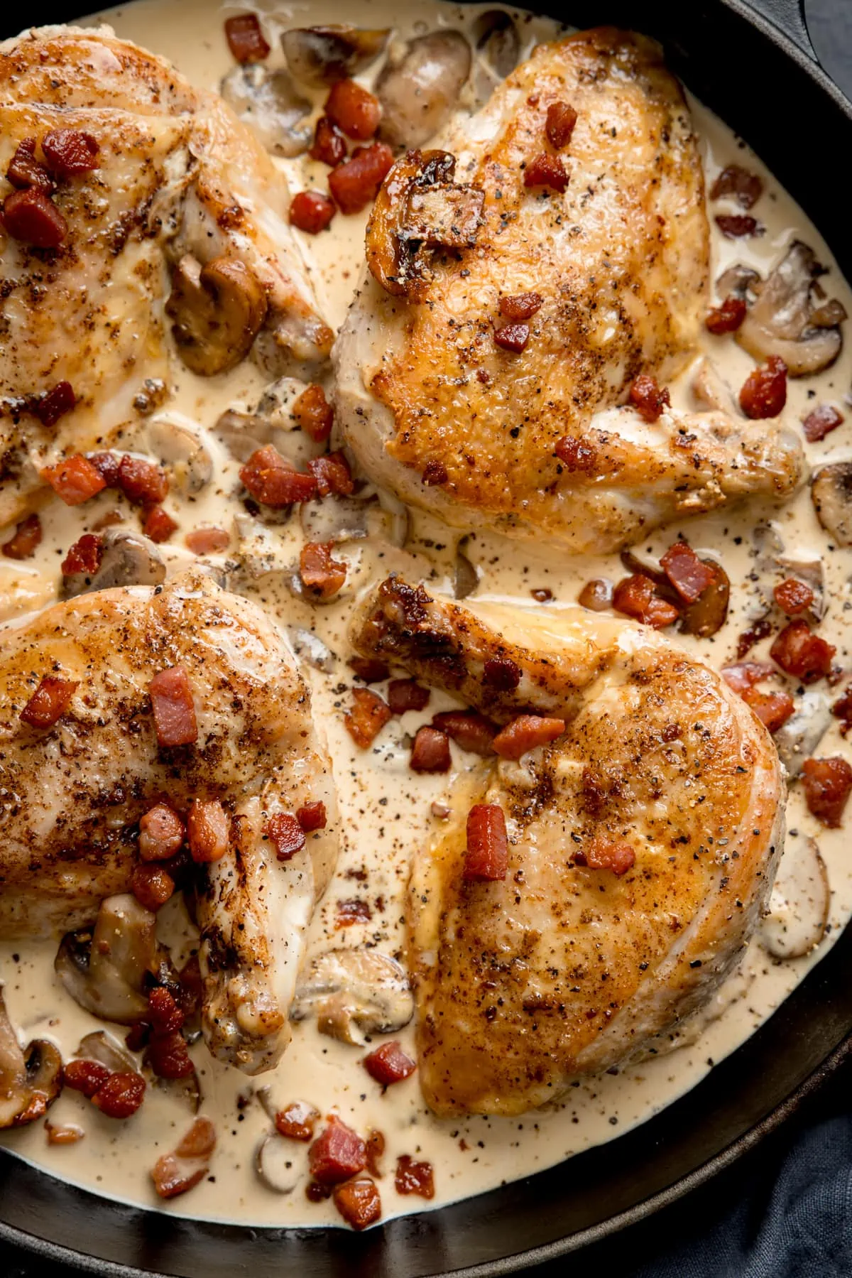 Overhead close-up image of chicken supreme with mushrooms and pancetta in a black pan. There are four chicken supremes in the pan.