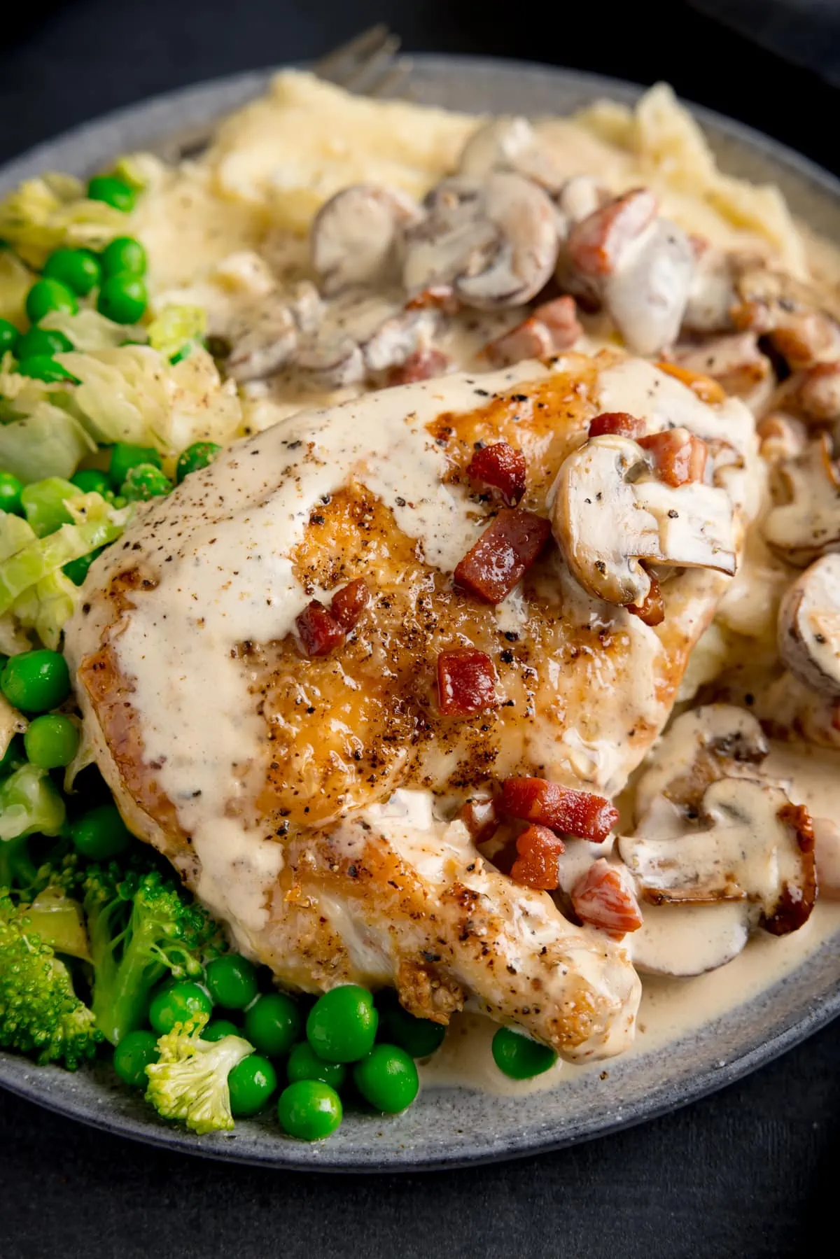 Chicken supreme with mushrooms and pancetta on a plate with mashed potato and green vegetables. The creamy sauce is drizzled over the chicken.