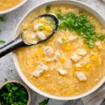 Overhead square shot of a white bowl filled with chicken and sweetcorn soup. The soup is topped with spring onions and there is a black spoon sticking out. There are some spring onions also in shot.