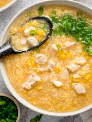 Overhead square shot of a white bowl filled with chicken and sweetcorn soup. The soup is topped with spring onions and there is a black spoon sticking out. There are some spring onions also in shot.