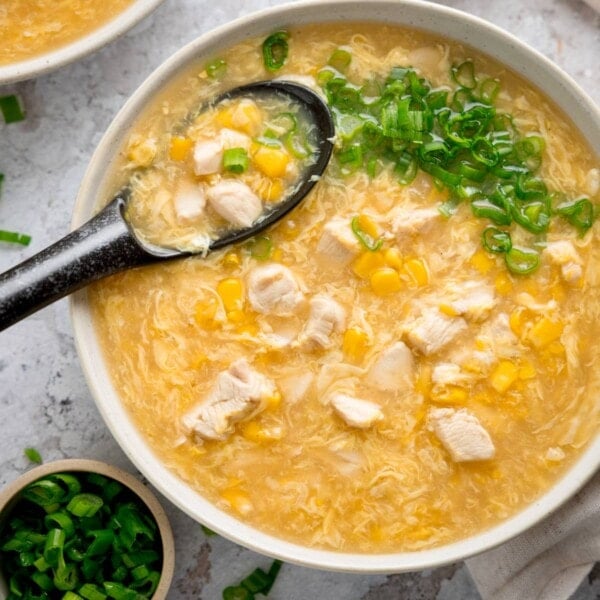 Overhead square shot of a white bowl filled with chicken and sweetcorn soup. The soup is topped with spring onions and there is a black spoon sticking out. There are some spring onions also in shot.