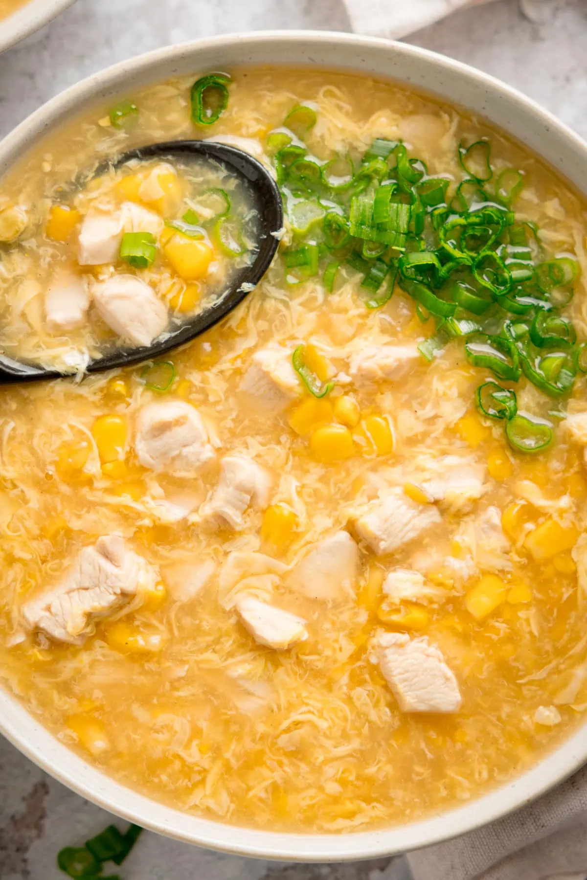 Close up overhead shot of a white bowl filled with chicken and sweetcorn soup. There are chopped spring onions on top and a black spoon sticking out of the soup.