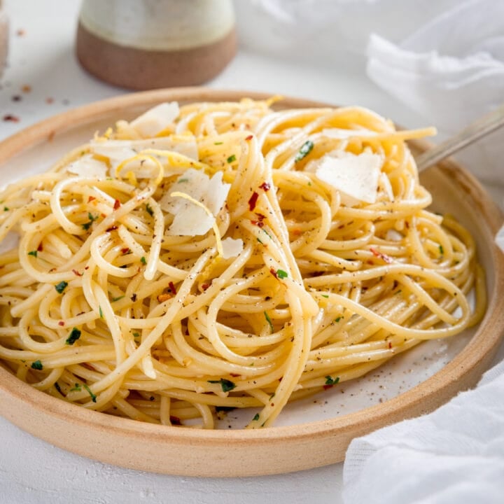 Square image of Spaghetti aglio e olio on white plate with stone rim. The plate is on a white background next to a white napkin. There is a little stone jug also in shot.