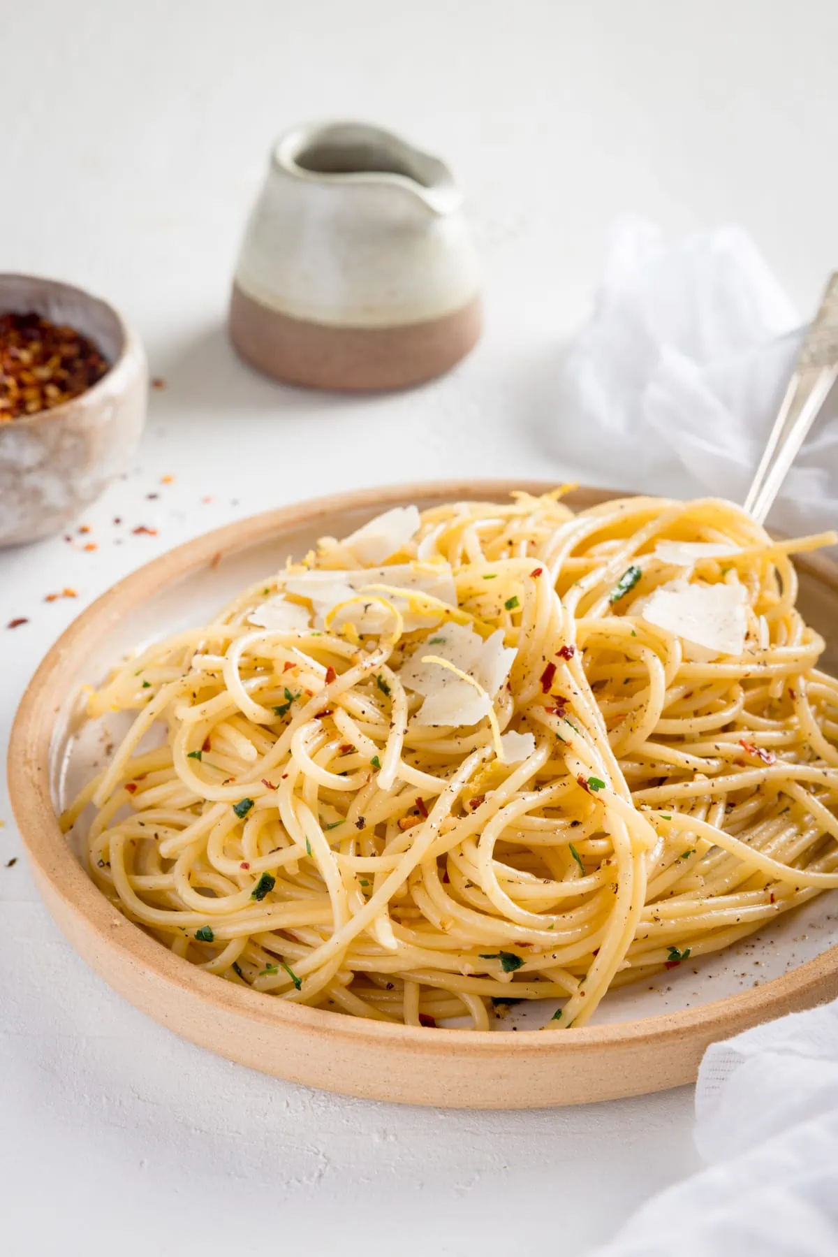Spaghetti aglio e olio on white plate with a stone rim. The plate is on a white background next to a white napkin. There is a pinch pot of chilli flakes and a little stone jug also in shot.