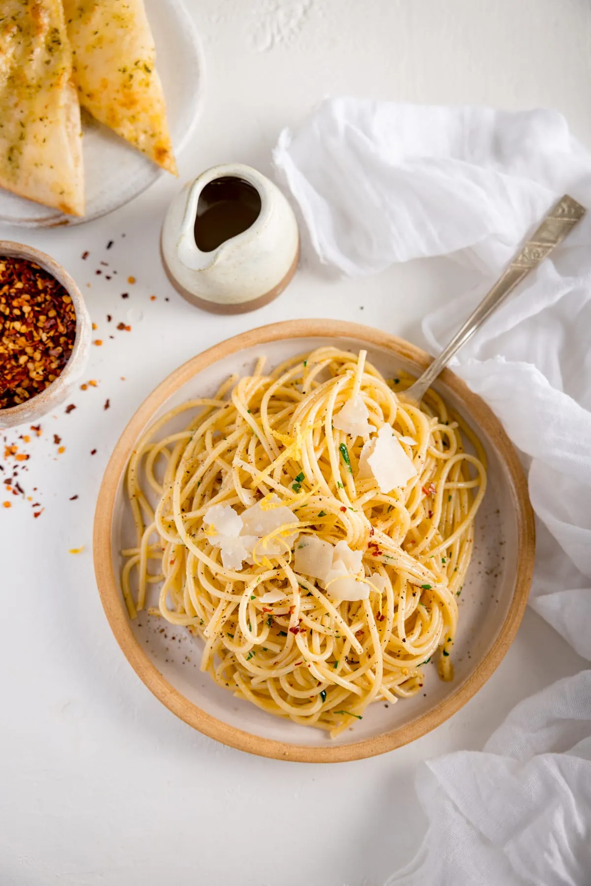Overhead shot of Spaghetti aglio e olio on white plate with a stone rim. The plate is on a white background next to a white napkin. There is a pinch pot of chilli flakes, a white plate with garlic bread slices and a little stone jug also in shot.