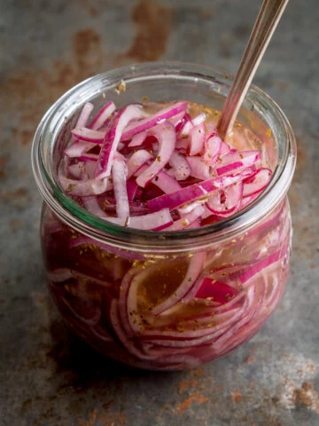Marinated sliced red onions in a glass jar with a metal spoon sticking out. The jar is on a vintage steel surface.