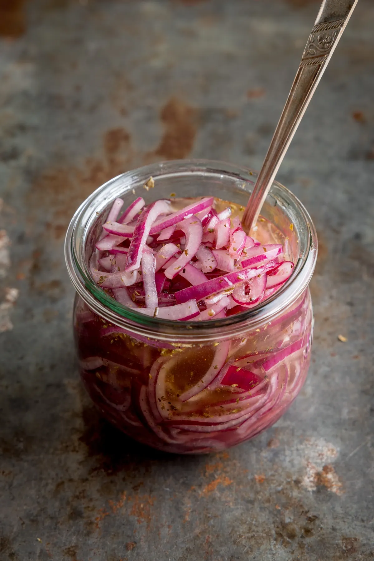 Marinated sliced red onions in a glass jar with a metal spoon sticking out. The jar is on a vintage steel surface.