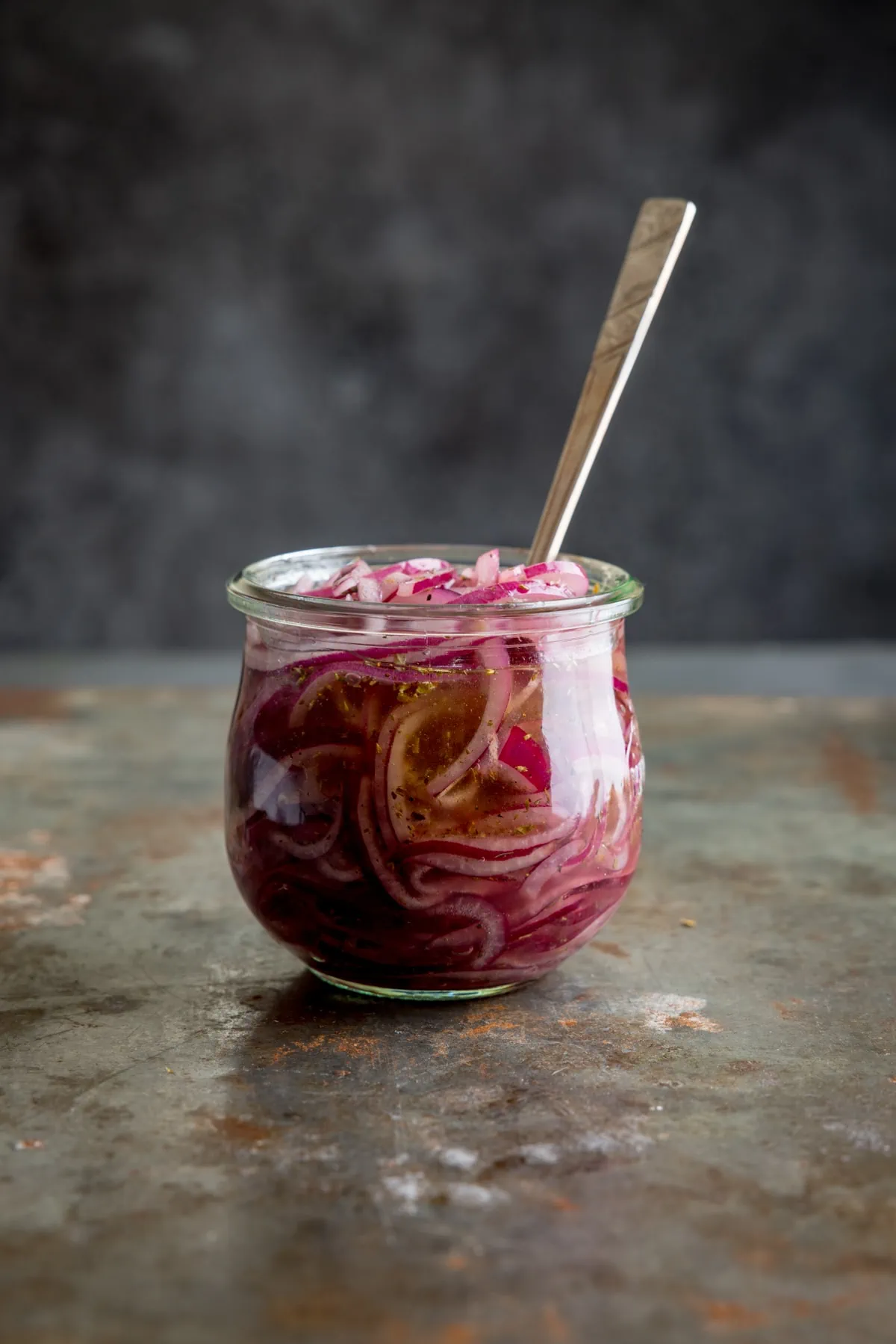 Side view of marinated, sliced red onions in a glass jar with a metal spoon sticking out. The glass stands on a vintage steel surface against a dark background.