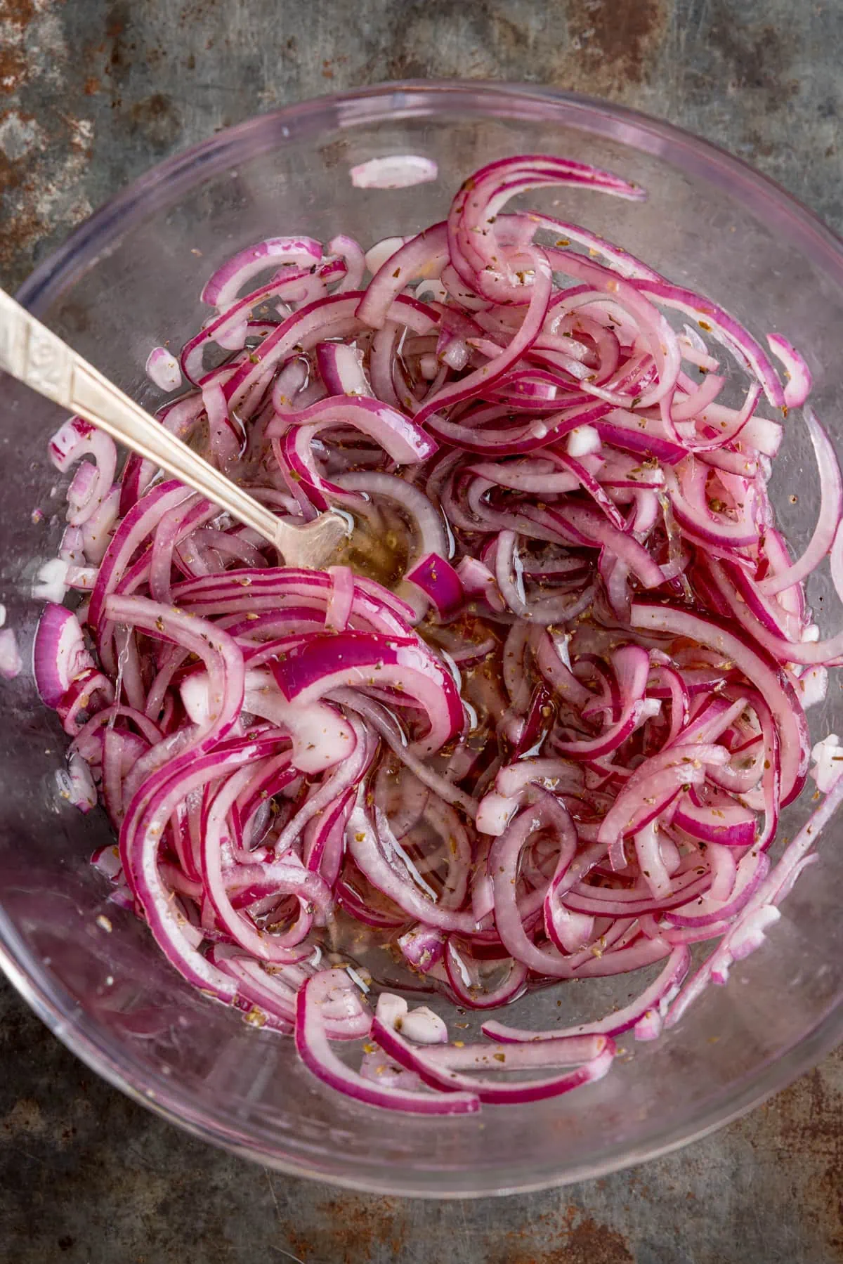 Overhead image of marinated sliced red onion in a glass bowl with a metal spoon sticking out.