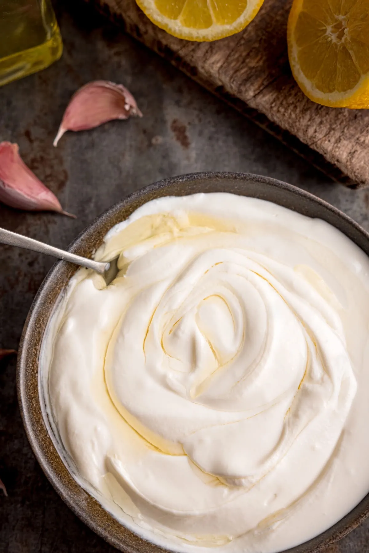 Overhead tall image of whipped feta dip in a dark bowl, swirled with olive oil. There is a spoon sticking out of the bowl. The bowl is on a dark background and there are ingredients scattered around.