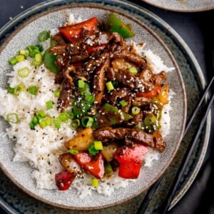 Beef in black bean sauce stir fry with boiled rice in a bowl, topped with spring onions and sesame seeds. There is a pair of black chopsticks next to the bowl.
