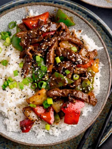 Beef in black bean sauce stir fry with boiled rice in a bowl, topped with spring onions and sesame seeds. There is a pair of black chopsticks next to the bowl.