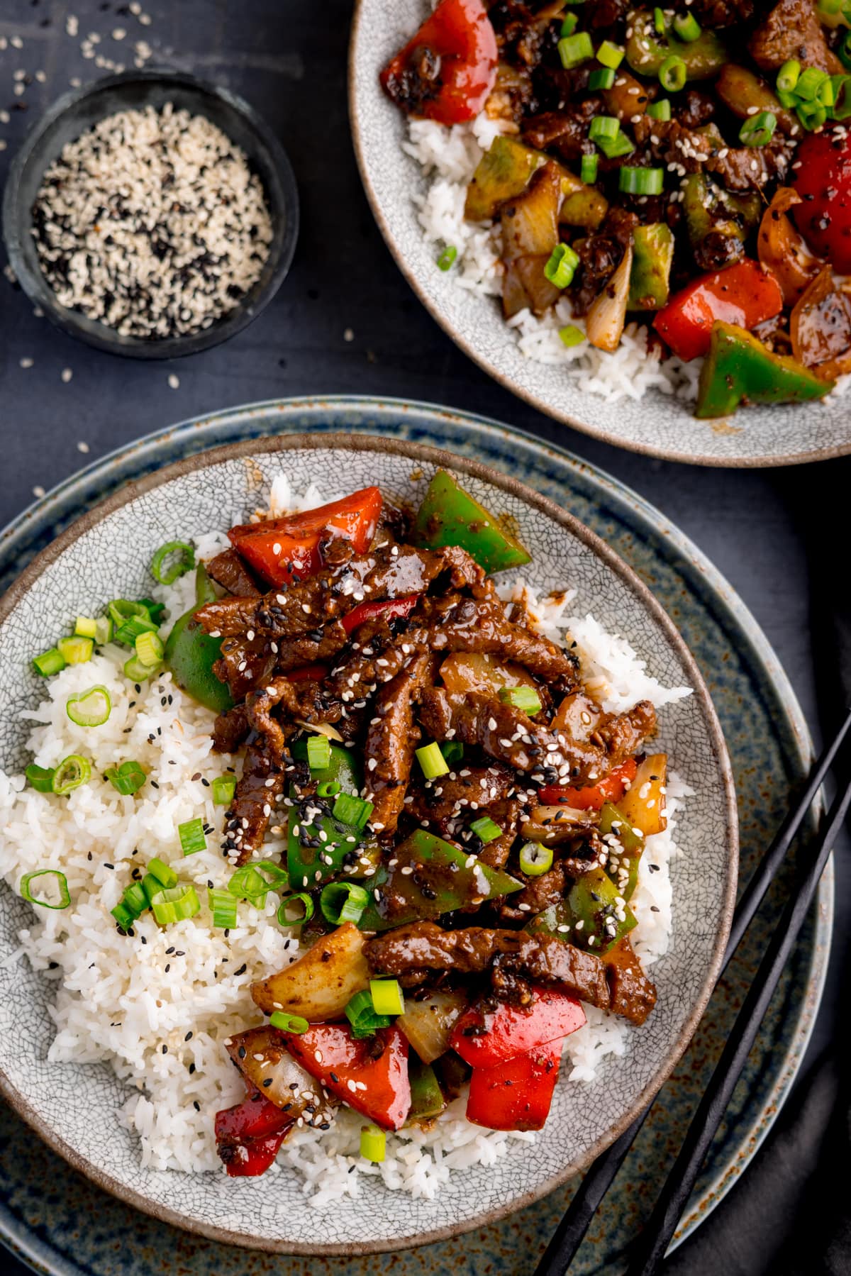 Beef in black bean sauce stir fry with boiled rice in a bowl, topped with spring onions and sesame seeds. The bowls is on a dark surface and there is a further bowl in shot. There is a small bowl of black and white sesame seeds nearby.