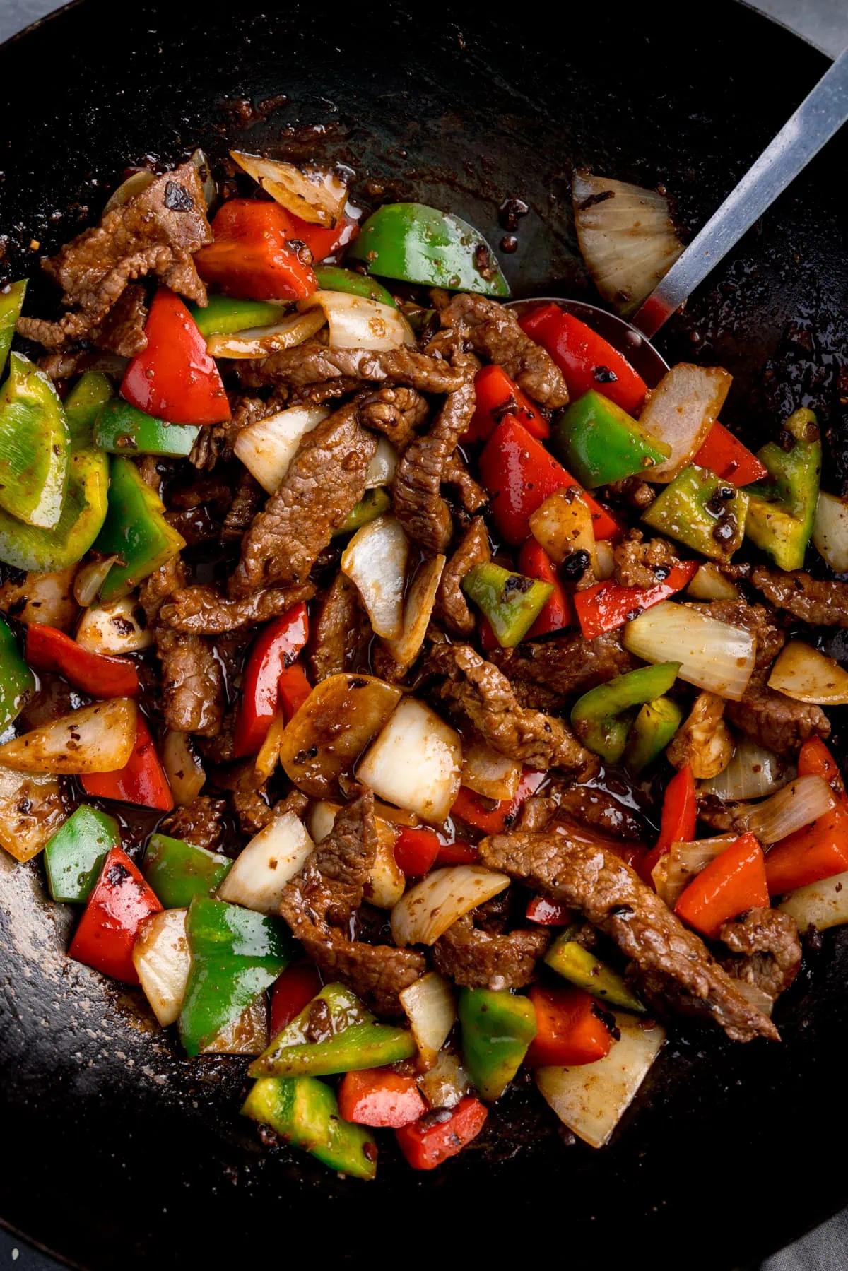 Close up overhead shot of beef in black bean sauce stir fry, with peppers and onions, in a wok, with a spatula sticking out.