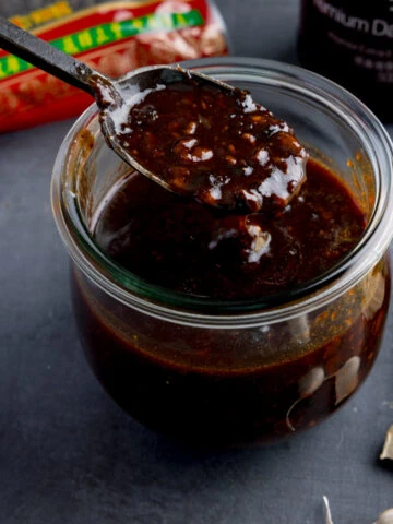 square image of a glass jar filled with homemade black bean sauce. There is a black spoon, taking a spoonful from the jar. The jar is on a dark surface and there are ingredients in the background