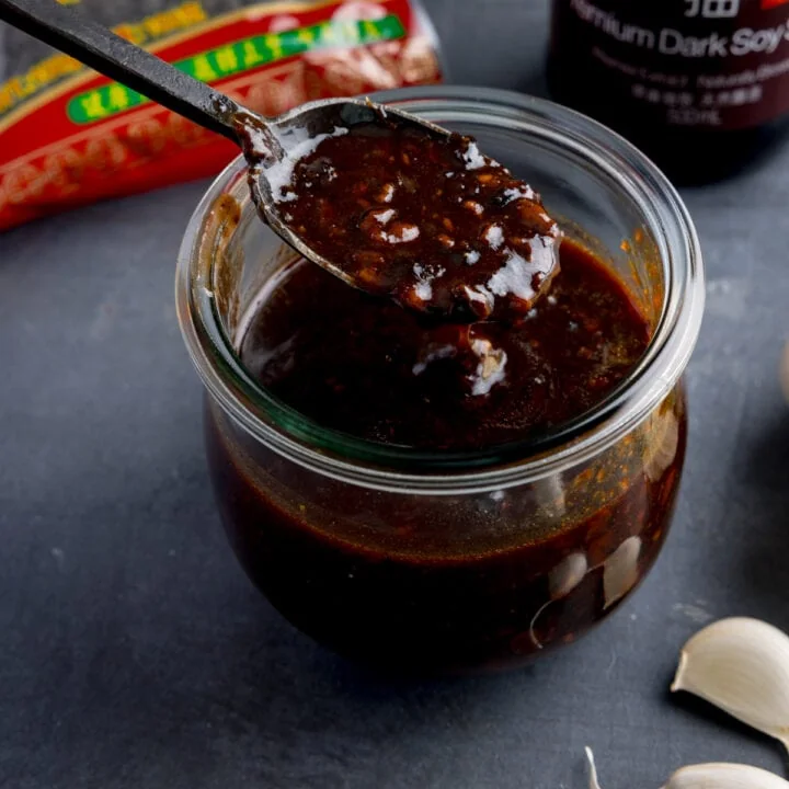 square image of a glass jar filled with homemade black bean sauce. There is a black spoon, taking a spoonful from the jar. The jar is on a dark surface and there are ingredients in the background