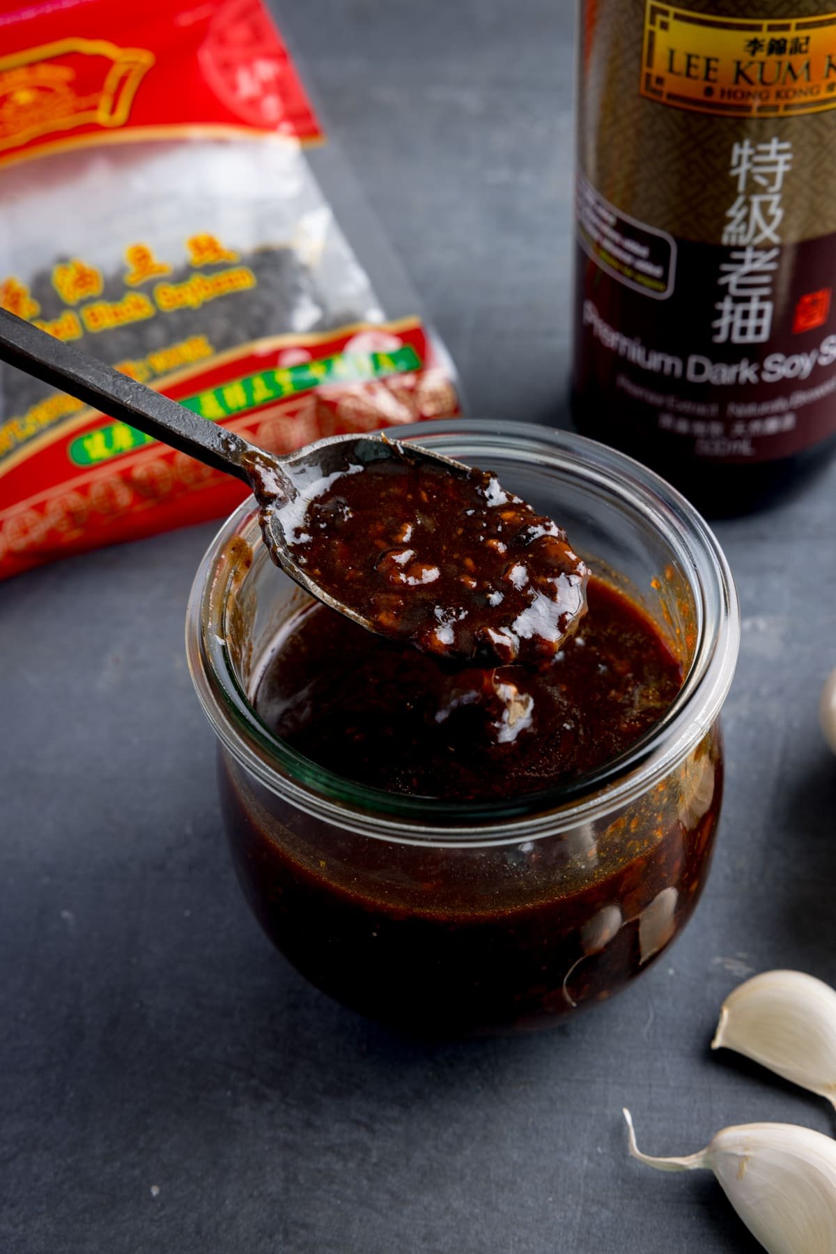 A glass jar filled with homemade black bean sauce. There is a black spoon, taking a spoonful from the jar.
The jar is on a dark surface and there are ingredients scattered around.