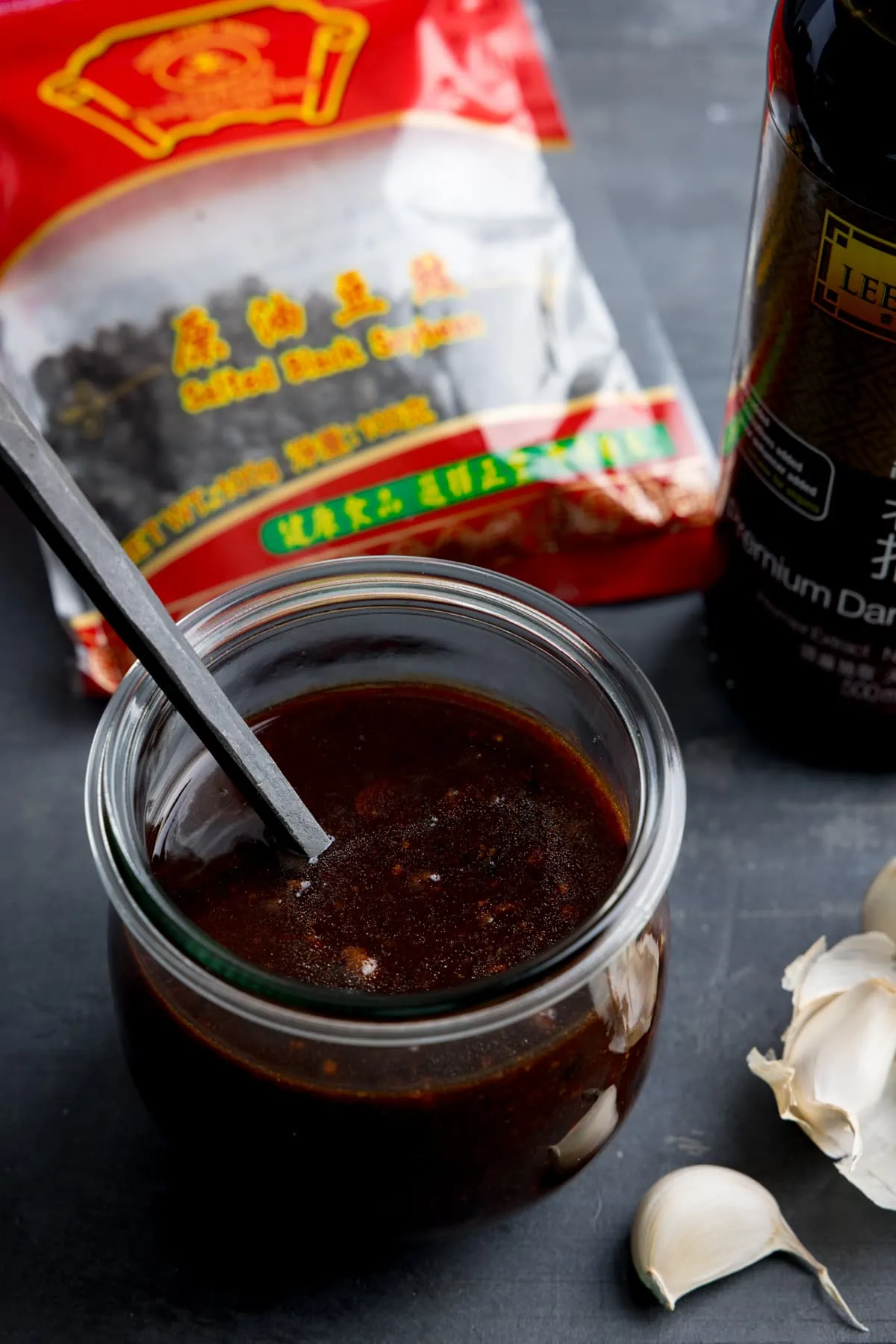 A glass jar filled with homemade black bean sauce. There is a black spoon sticking out of the jar. The jar is on a dark surface and there are ingredients in the background
