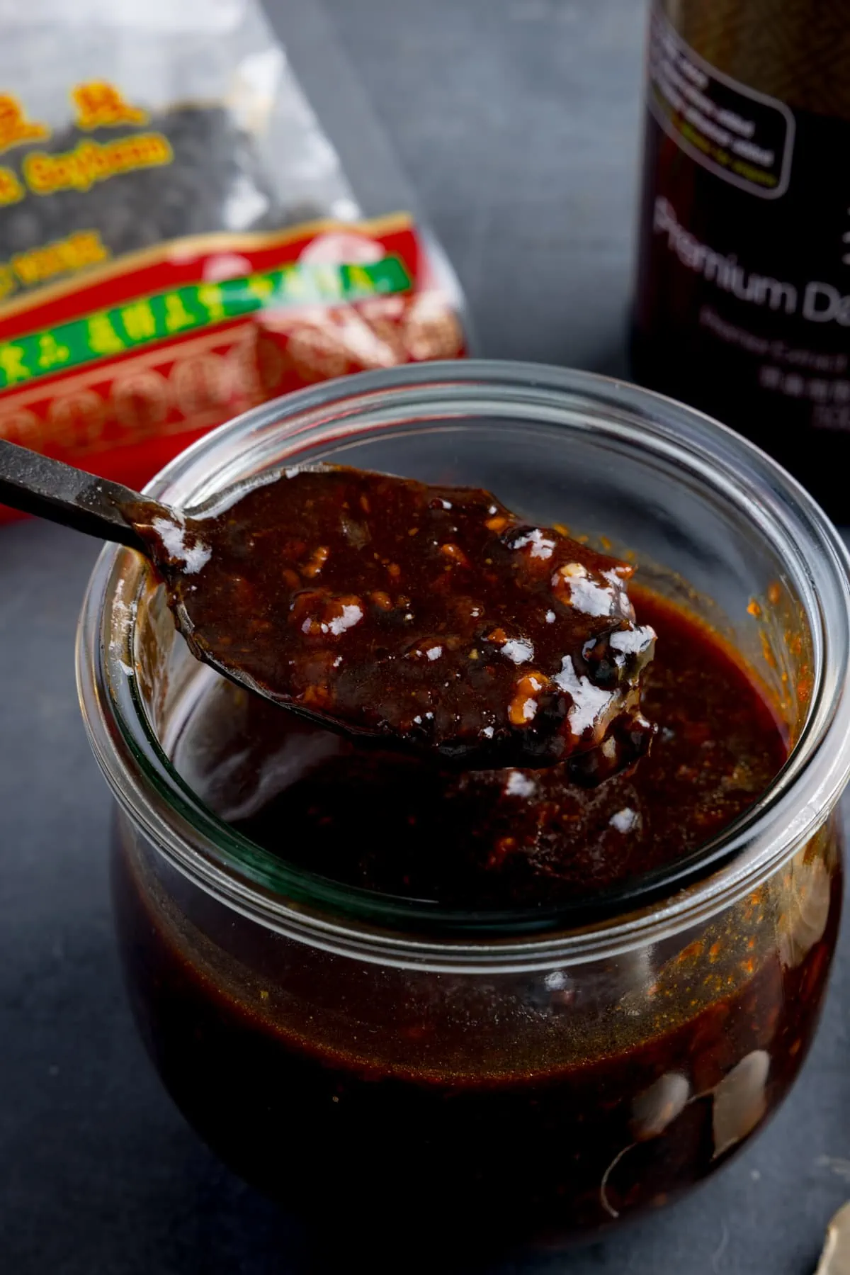 A glass jar filled with homemade black bean sauce. There is a black spoon, taking a spoonful from the jar. The jar is on a dark surface and there are ingredients in the background.