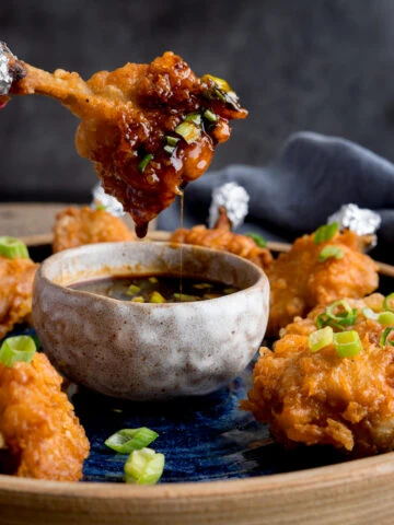 Square image of a plate of chicken lollipops with a bowl of spicy dipping sauce. One of the wings is being dipped into the sauce and is dripping. The wing is being held by fingers with pink-painted fingernails.