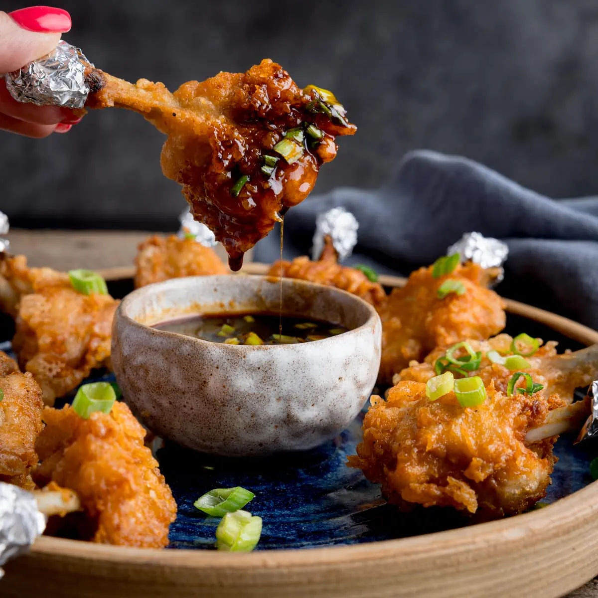 Square image of a plate of chicken lollipops with a bowl of spicy dipping sauce. One of the wings is being dipped into the sauce and is dripping. The wing is being held by fingers with pink-painted fingernails.