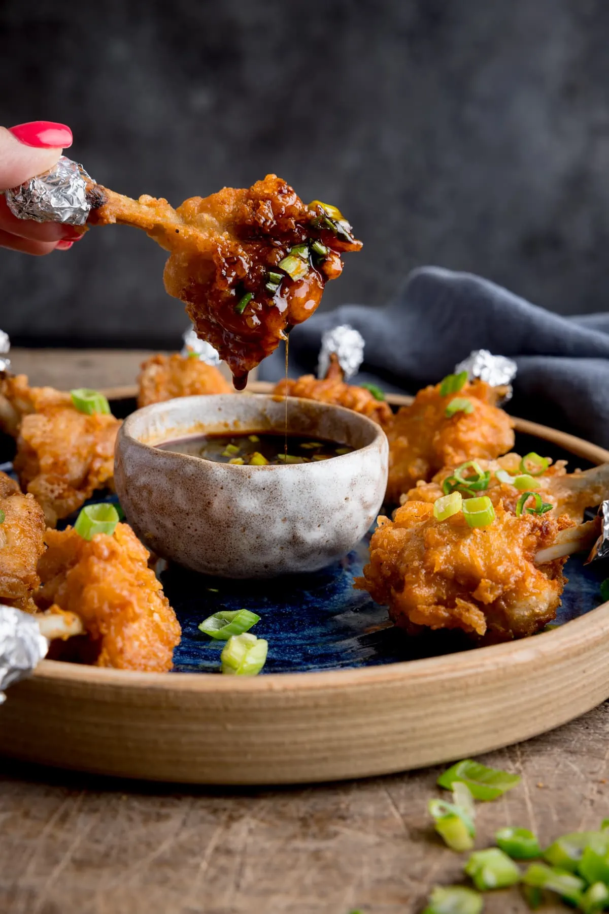 A plate of chicken lollipops with a bowl of spicy dipping sauce. One of the wings is being dipped into the sauce and is dripping. The wing is being held by fingers with pink-painted fingernails.