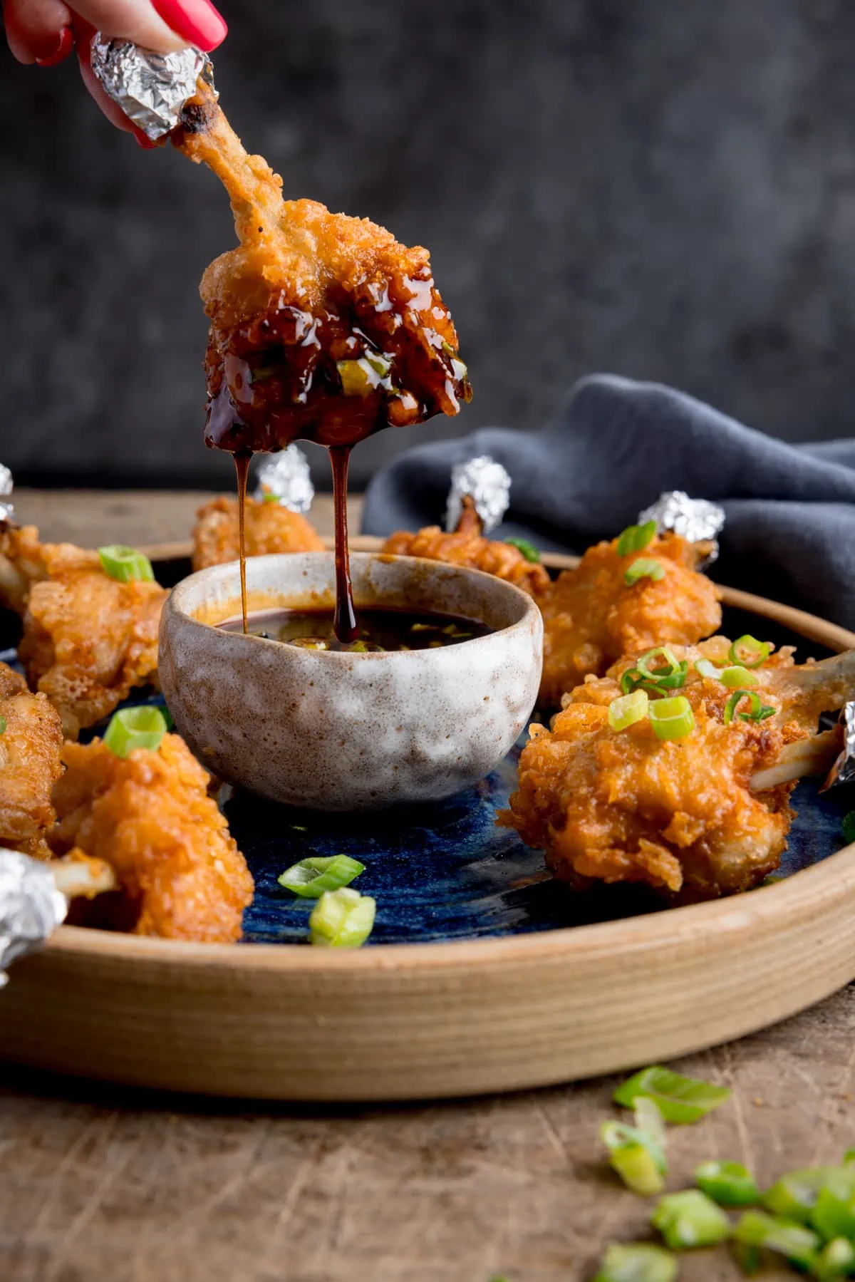 A plate of chicken lollipops with a bowl of spicy dipping sauce. on a wooden table. One of the wings is being dipped into the sauce and is dripping. The wing is being held by fingers with pink-painted fingernails.