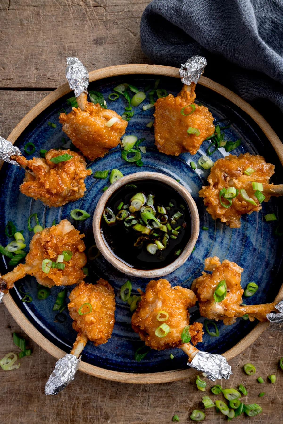 Chicken lollipops arranged in a circle on a blue plate with a dipping bowl of sticky spicy sauce int he middle. The lollipops and sauce have been sprinkled with chopped spring onions and the plate is in a wooden table next to a blue napkin.