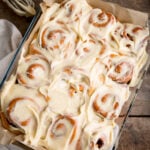 Overhead square image of 12 cinnamon rolls in a baking dish, topped with cream cheese frosting. The dish is on a wooden table, next to a beige napkin. There is a whisk with cream cheese on nearby.