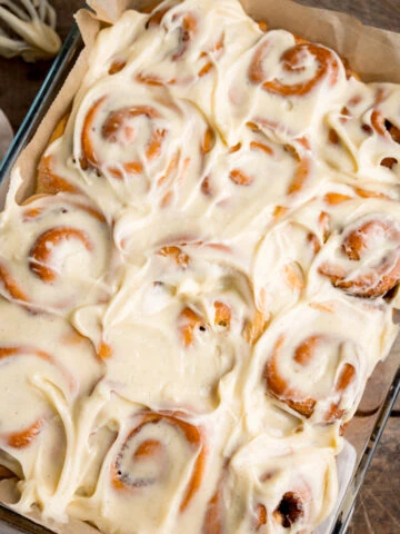 Overhead square image of 12 cinnamon rolls in a baking dish, topped with cream cheese frosting. The dish is on a wooden table, next to a beige napkin. There is a whisk with cream cheese on nearby.