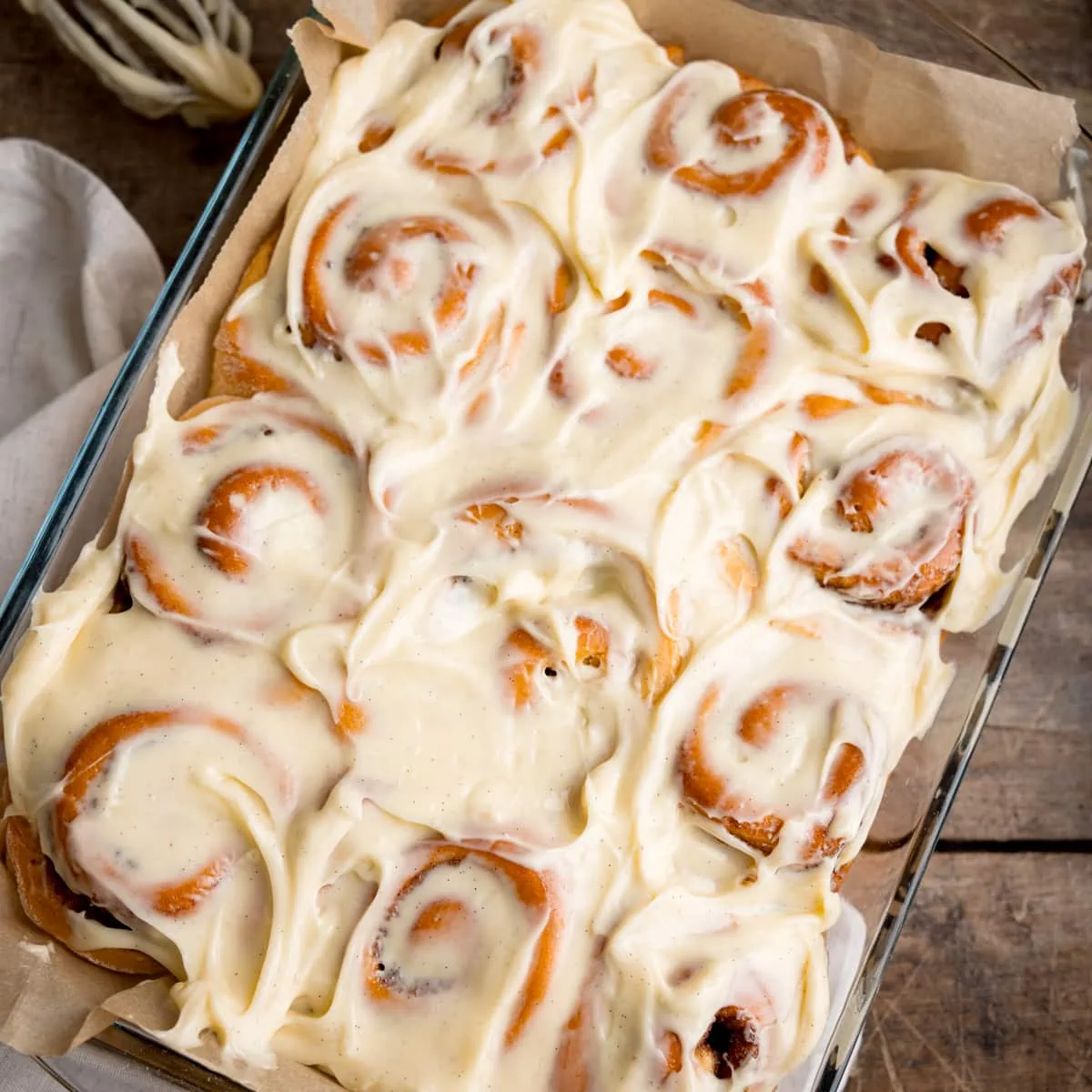 Overhead square image of 12 cinnamon rolls in a baking dish, topped with cream cheese frosting. The dish is on a wooden table, next to a beige napkin. There is a whisk with cream cheese on nearby.