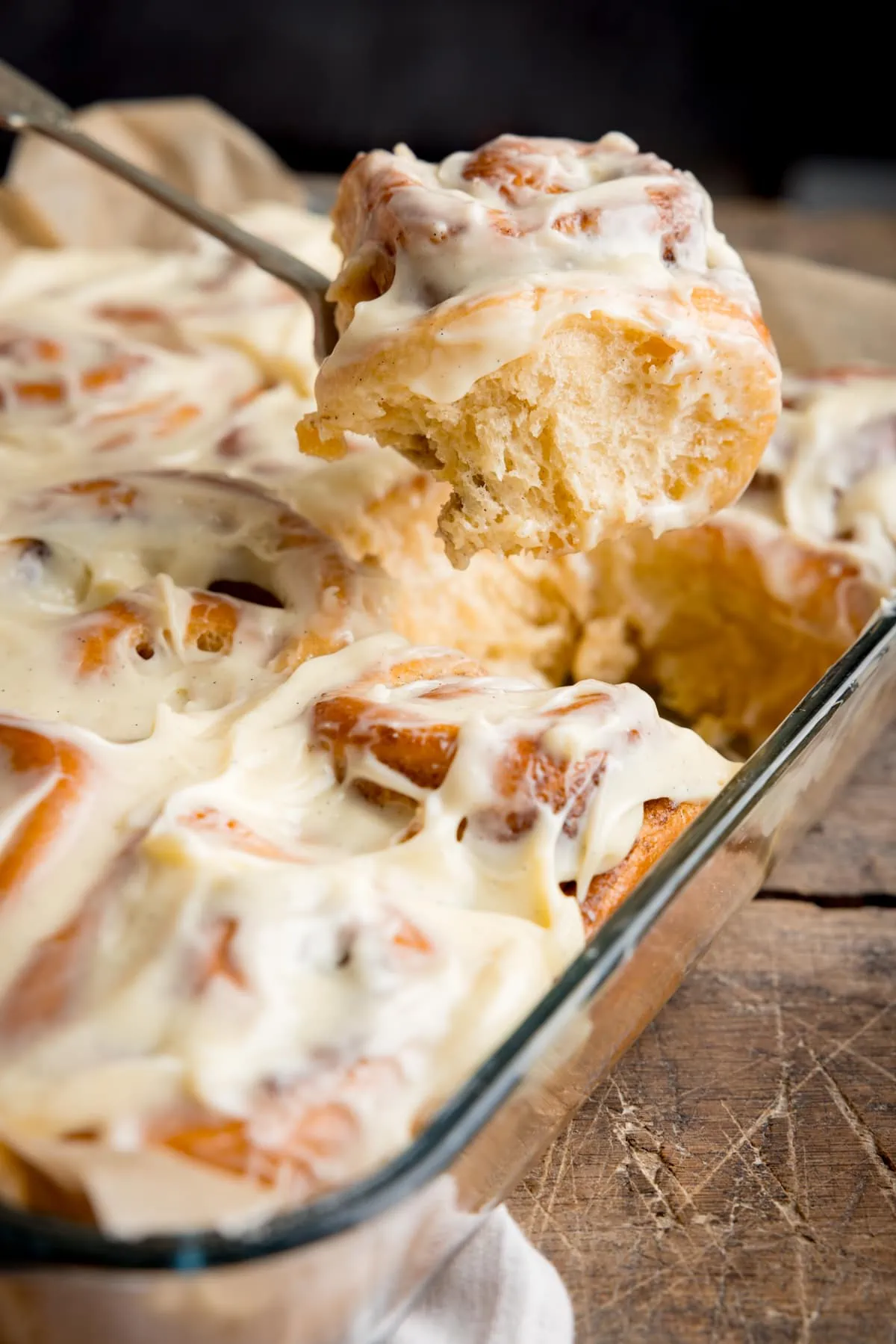 A glass baking dish of frosted cinnamon rolls with one cinnamon roll being taken out on a fork. The dish is on a wooden table and there is part of a beige napkin in shot.