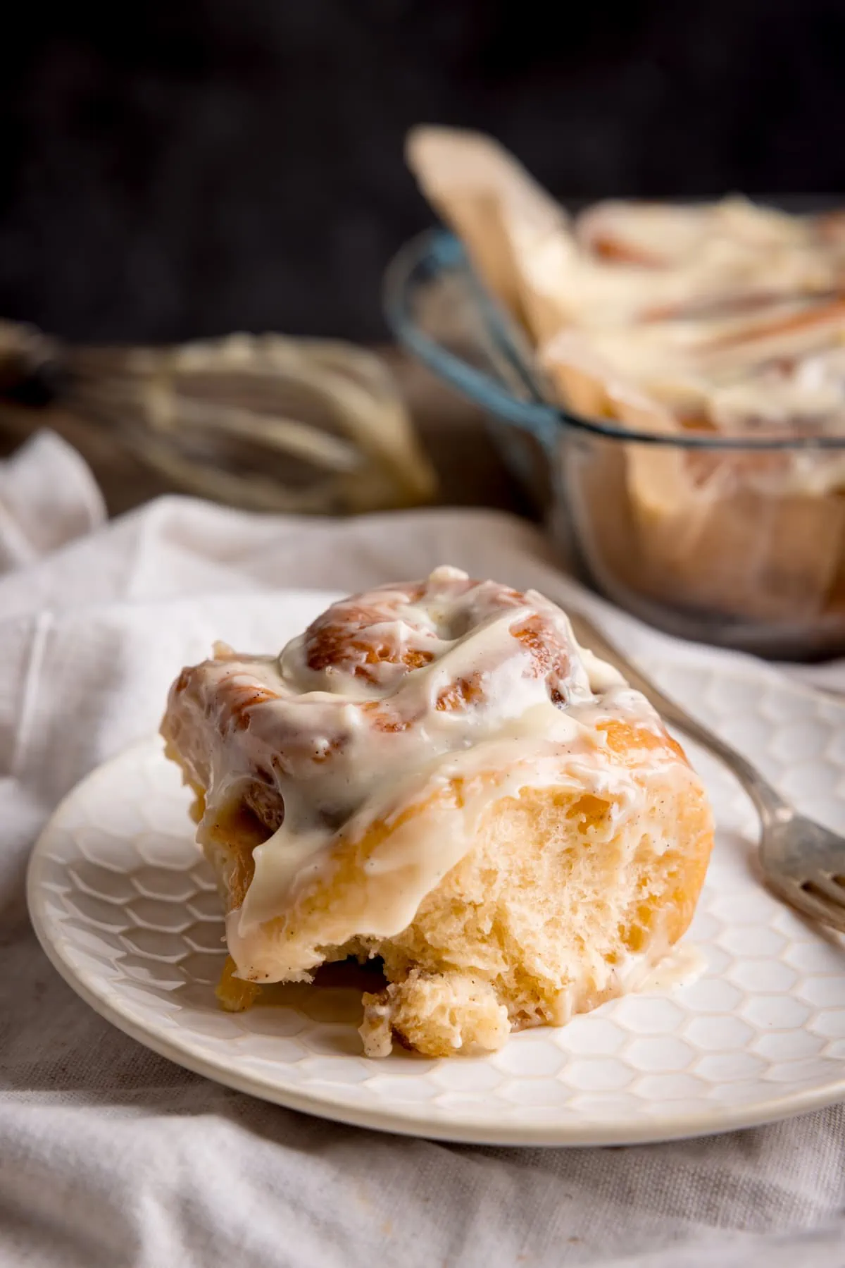 A single frosted cinnamon roll on a white plate with a small fork next to it. The plate is on a beige napkin. In the background there is a glass dish filled with cinnamon rolls and a whisk with cream cheese frosting on it.