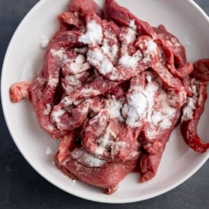 Overhead square image of a white bowl on a grey surface, filled with strips of raw beef steak, coated in bicarbonate of soda.