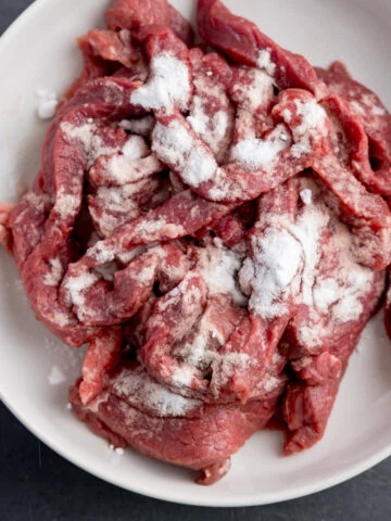 Overhead square image of a white bowl on a grey surface, filled with strips of raw beef steak, coated in bicarbonate of soda.