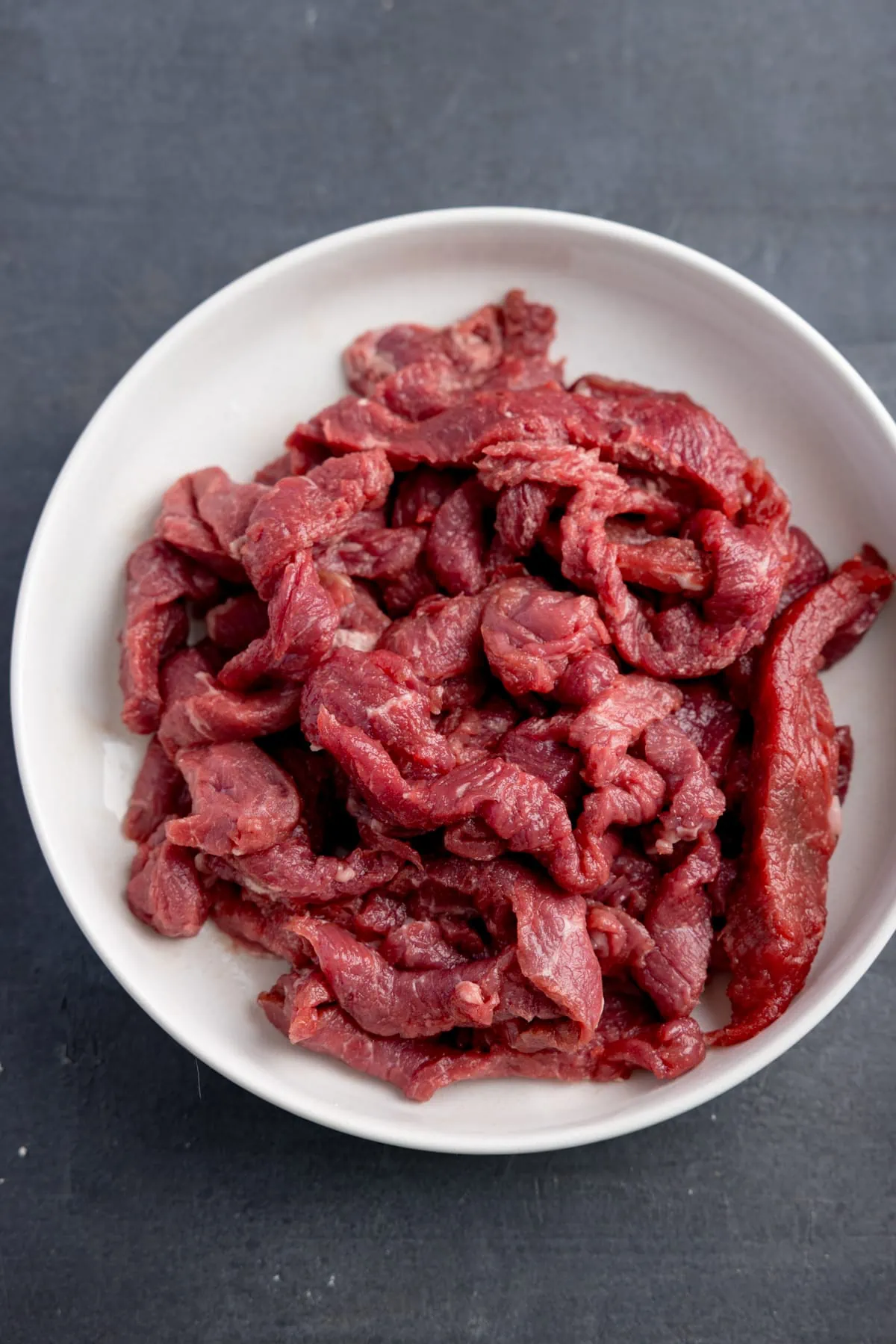 Overhead image of a white bowl on a grey surface, filled with strips of raw beef steak.